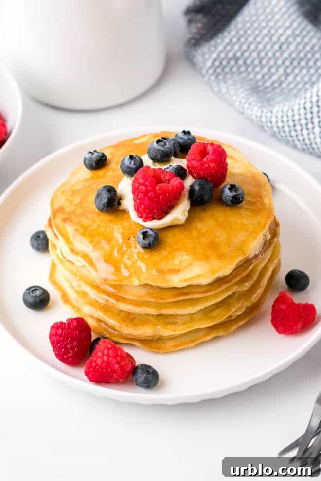 a stack of four fluffy homemade pancakes on a white plate with butter, syrup, blueberries and raspberries