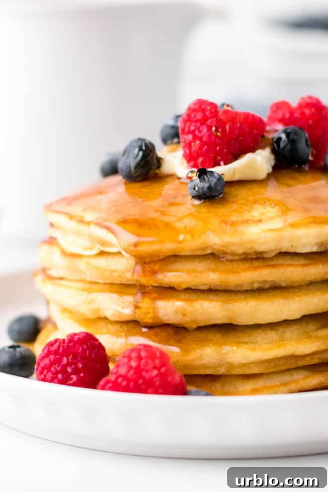 a stack of fluffy homemade pancakes on a white plate with butter, syrup and berries