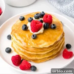 a stack of four fluffy homemade pancakes on a white plate with butter, syrup, blueberries and raspberries