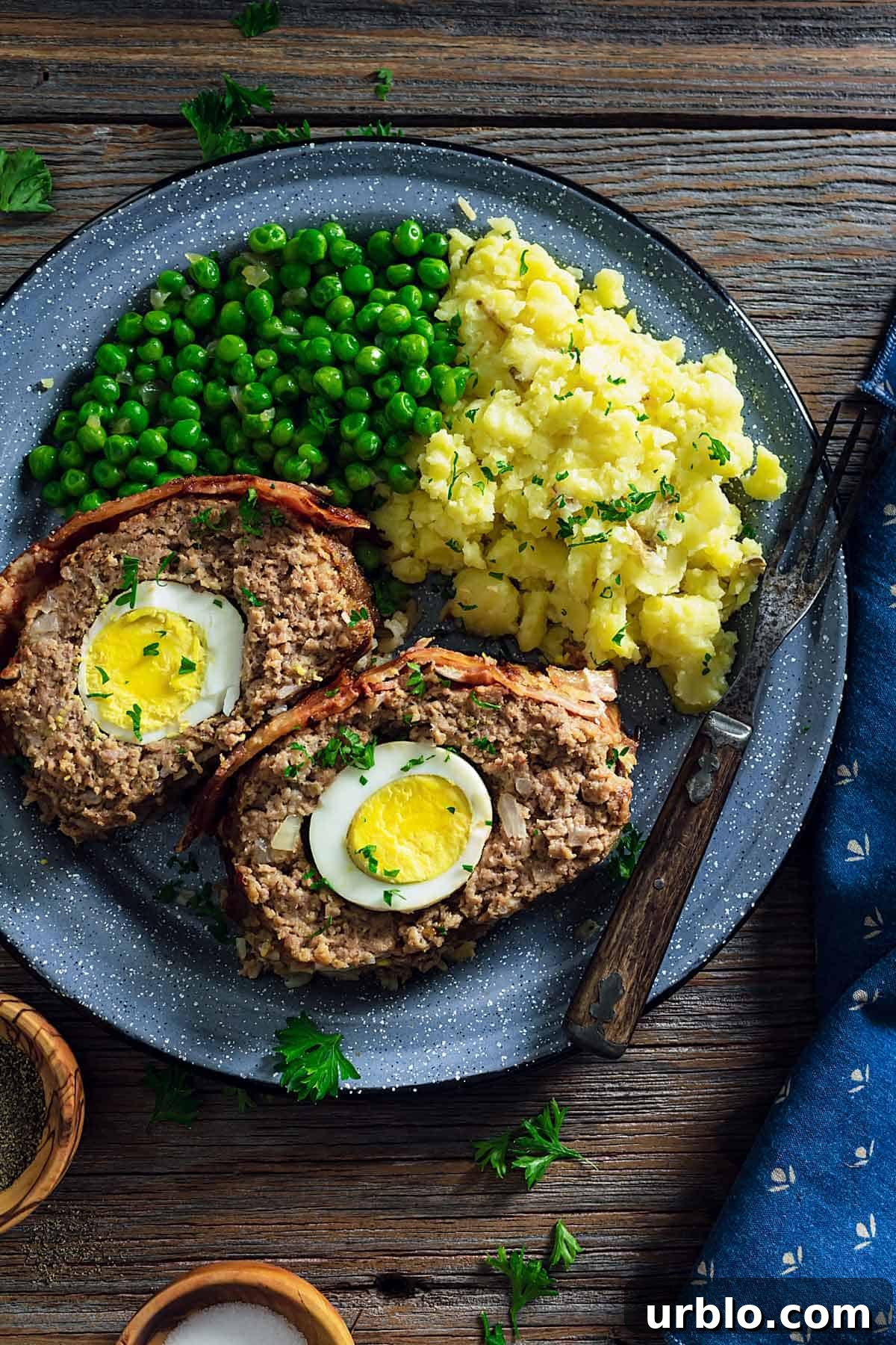 An overhead photo of two meatloaf slices showing the boiled eggs inside on a plate with mashed potatoes and green peas.