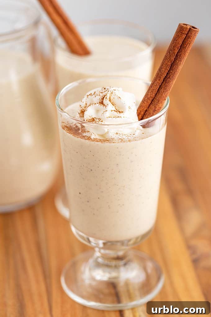 Eggnog in a glass with whipped cream, cinnamon stick, and a sprinkle of cinnamon on a wood surface
