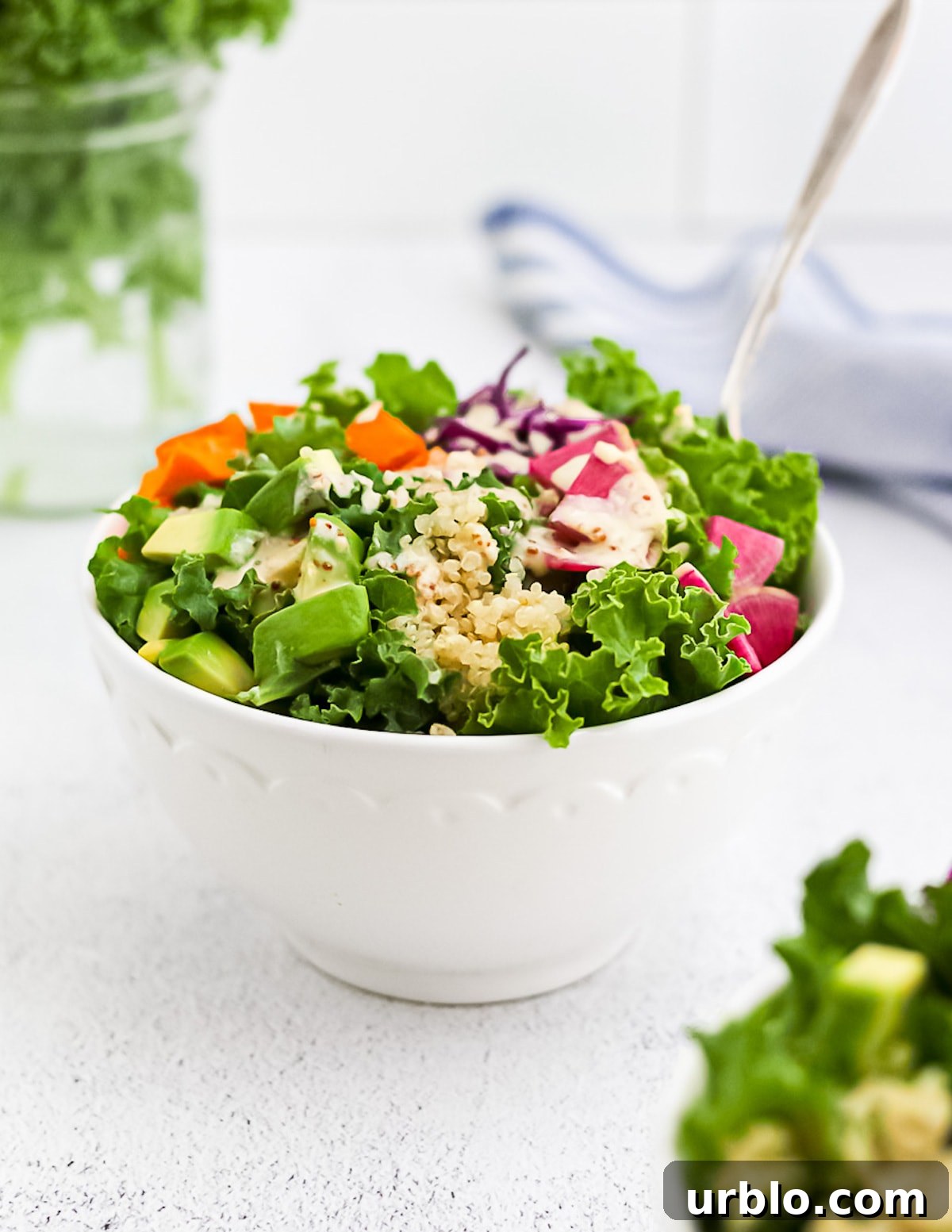 A kale salad with tahini dressing in a white bowl, containing radishes, sweet potatoes, cabbage, and avocado.