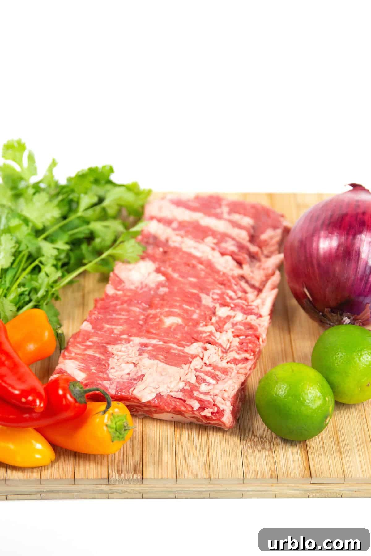 wood cutting board of raw flank steak, whole onion, peppers, cilantro, and lime