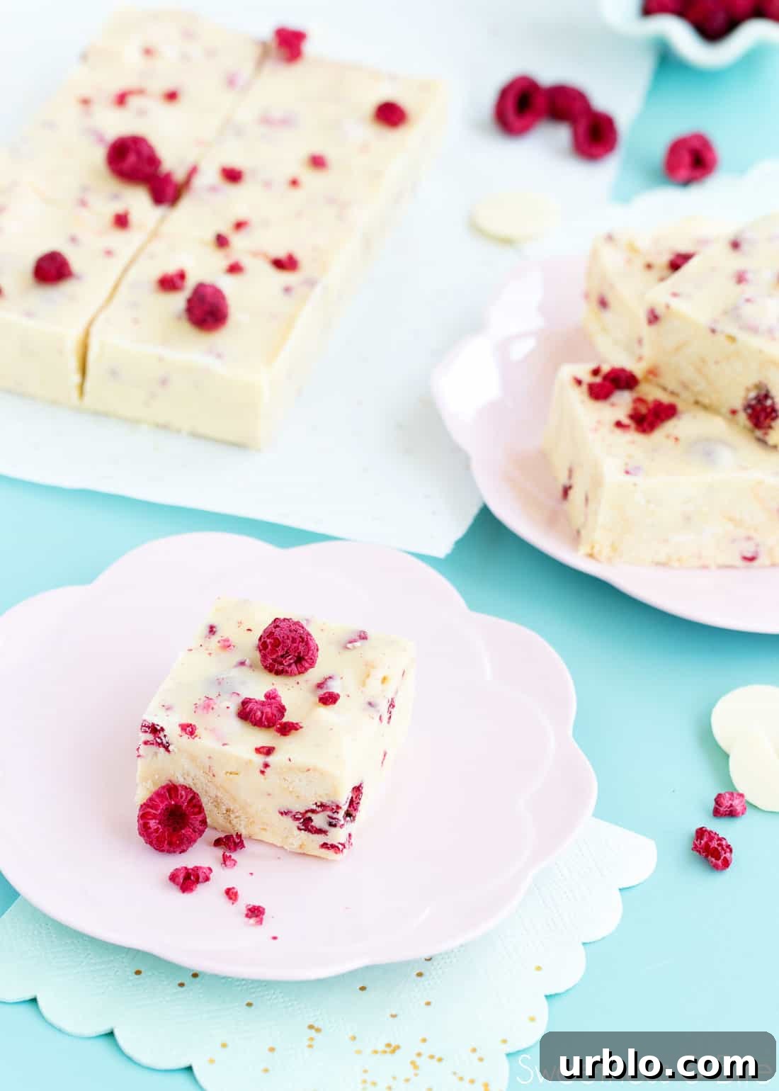 One piece of white choc raspberry slice on a pink flower shaped plate in the foreground. Other pieces of slice on a piece of parchment in the background, on a blue-green tabletop.