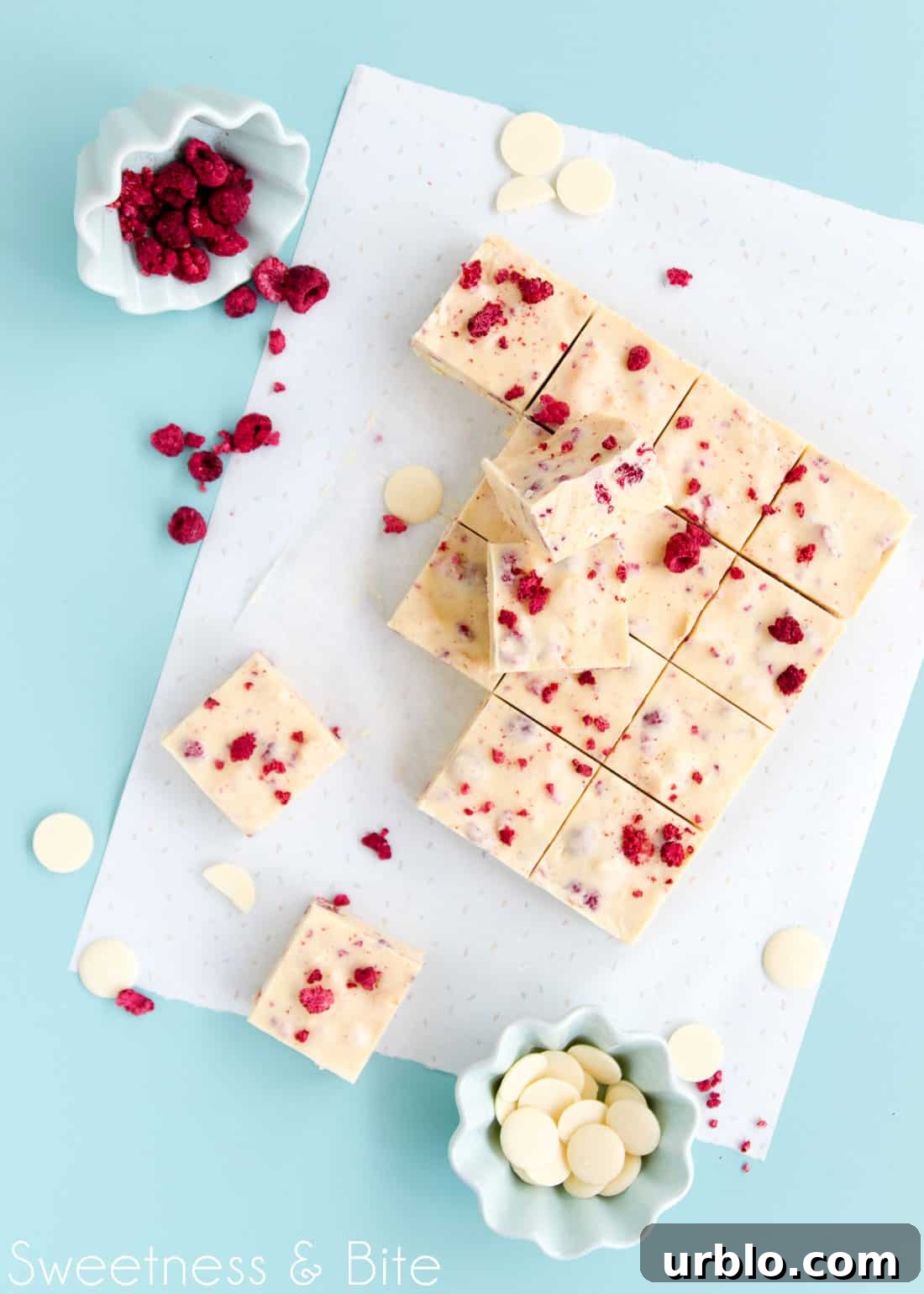 Two pieces of slice, sitting on top of the remaining pieces of slice below. A small green bowl of freeze-dried raspberry pieces sits on its side, with scattered pieces of raspberry falling out. White chocolate buttons and broken freeze-dried raspberries are scattered across the blue-green tabletop.