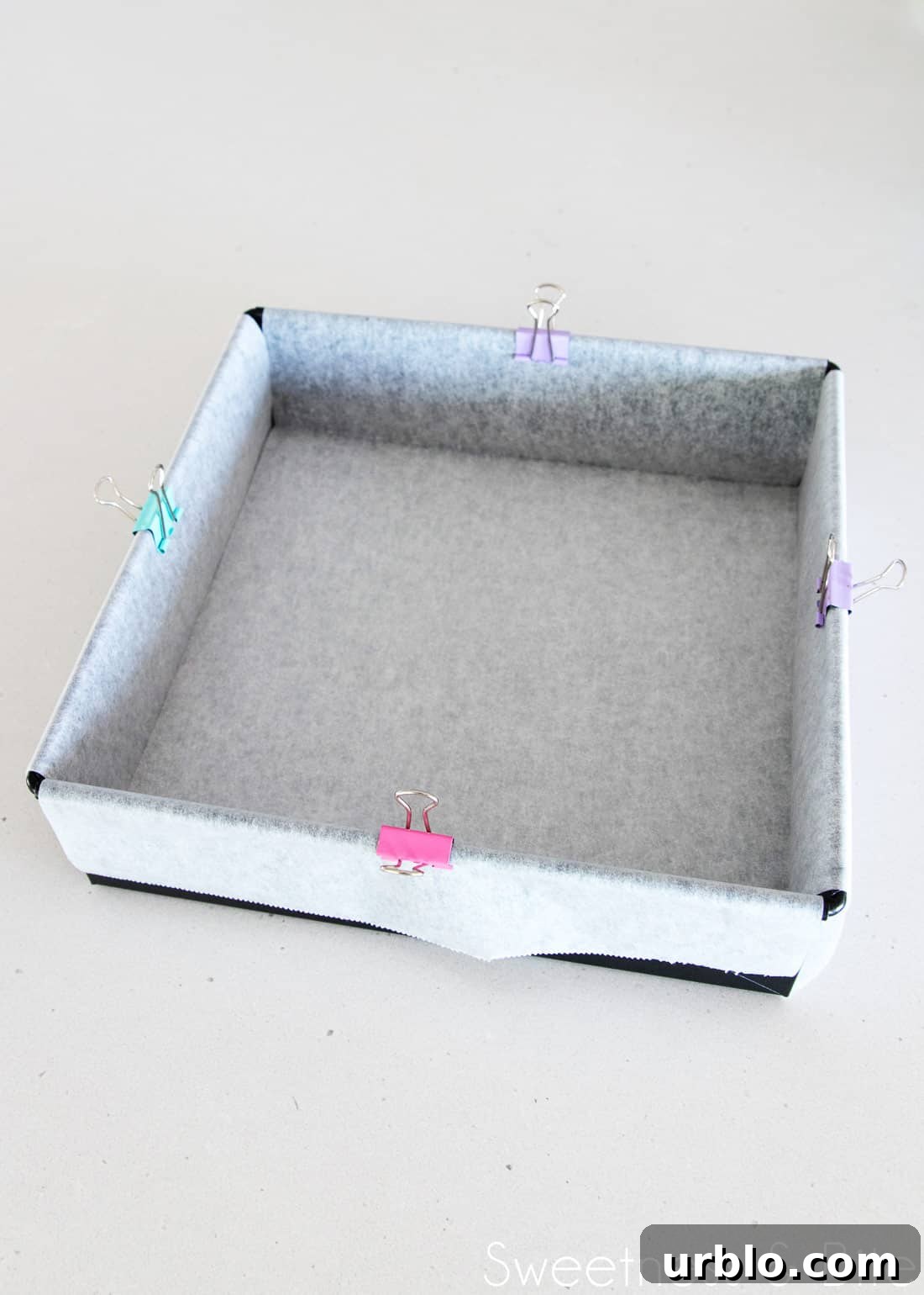 A dark metal square cake pan, with two strips of baking paper criss-crossed to line the pan, with coloured clips on each edge to hold the paper in place.