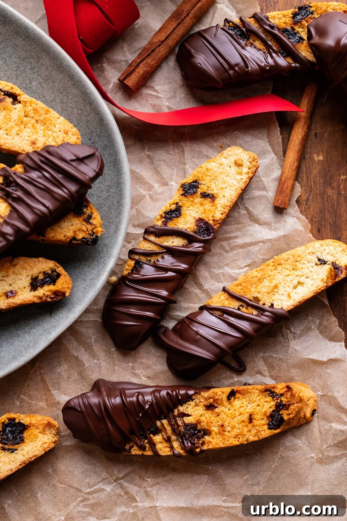 Chocolate dipped biscottis arranged on parchment paper, with a plate of biscottis next to them. Around the biscottis are red ribbon and cinnamon sticks.