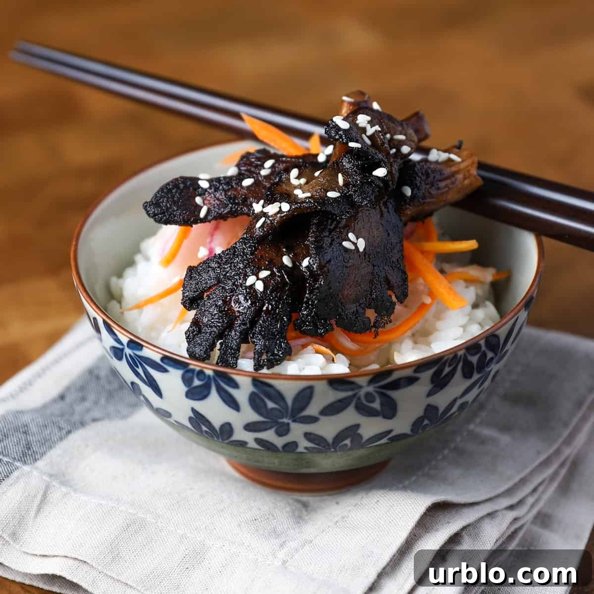 Photo of a pretty bowl filled with sushi rice, pickled salad and Char Siu mushrooms, set on a napkin with chop sticks resting.