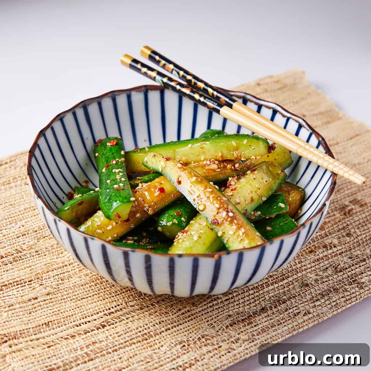Photo of a blue and white striped bowl filled with Chinese pickled cucumbers with chop sticks balancing on top of the bowl and a bamboo mat underneath.