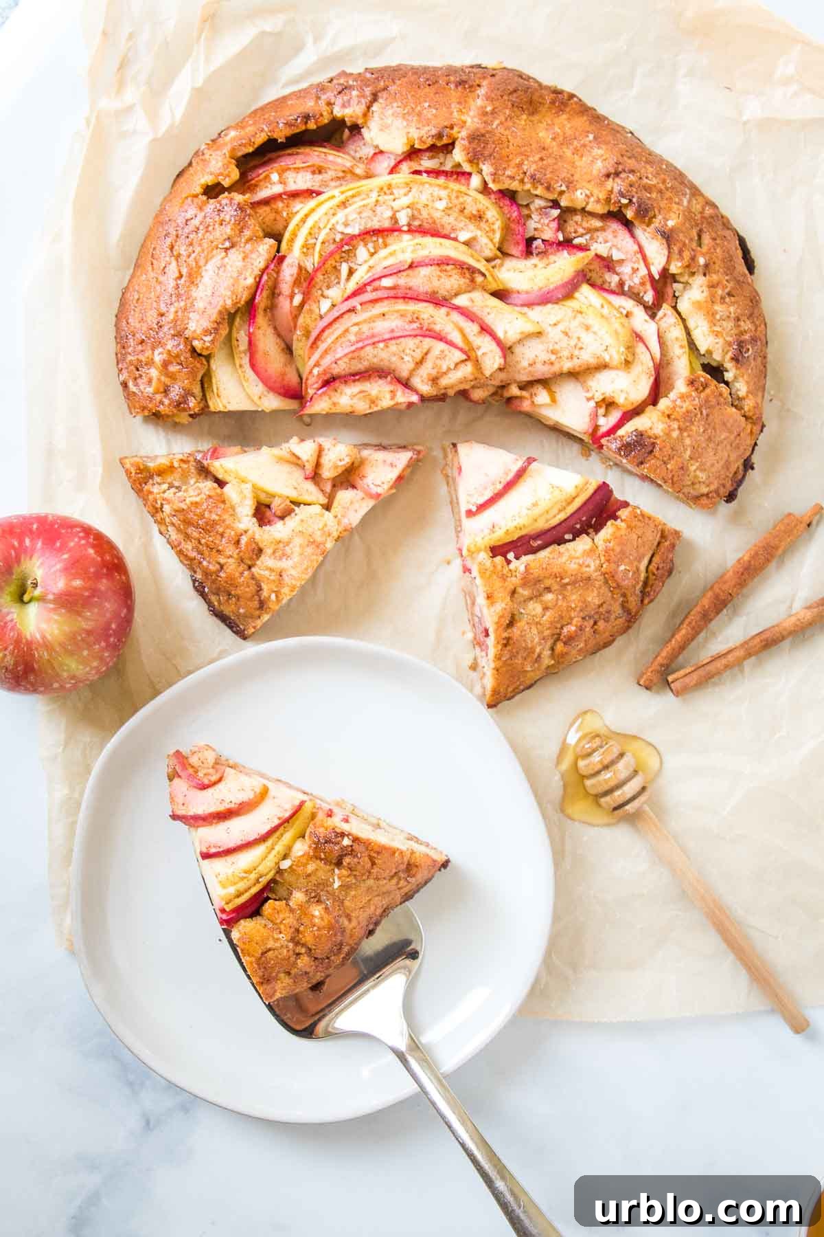 Free form apple galette, half of it sliced. One piece is on a plate. There is an apple, some cinnamon sticks and a dollop of honey on the parchment paper that the galette is also on.