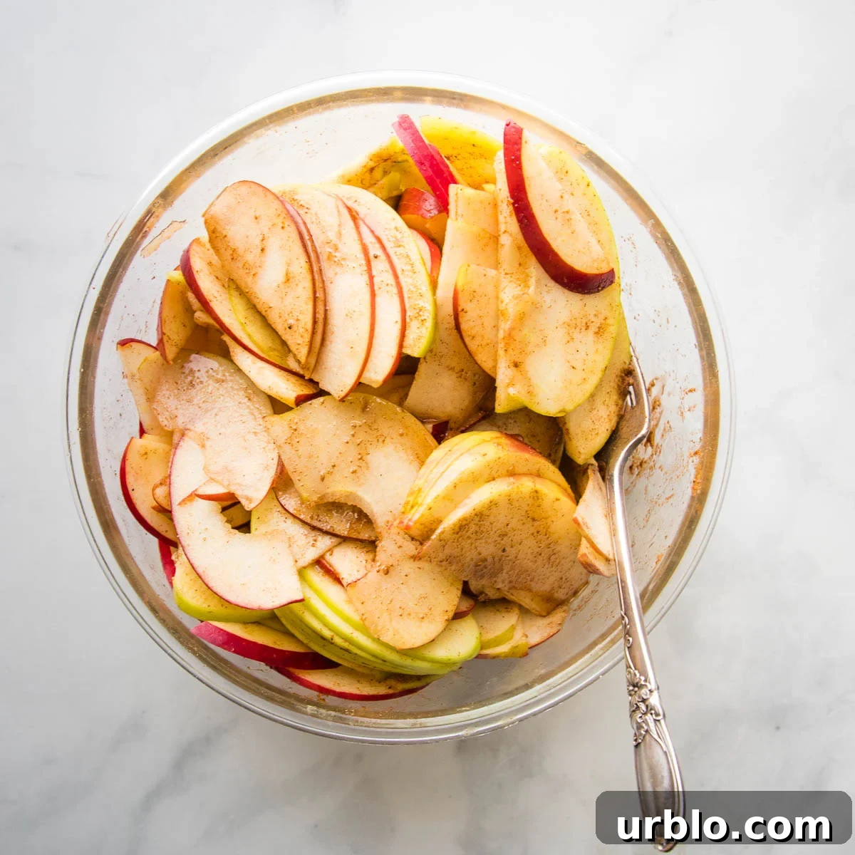 Green and Red Sliced Apples, mixed with apple pie spices, in a large glass bowl.