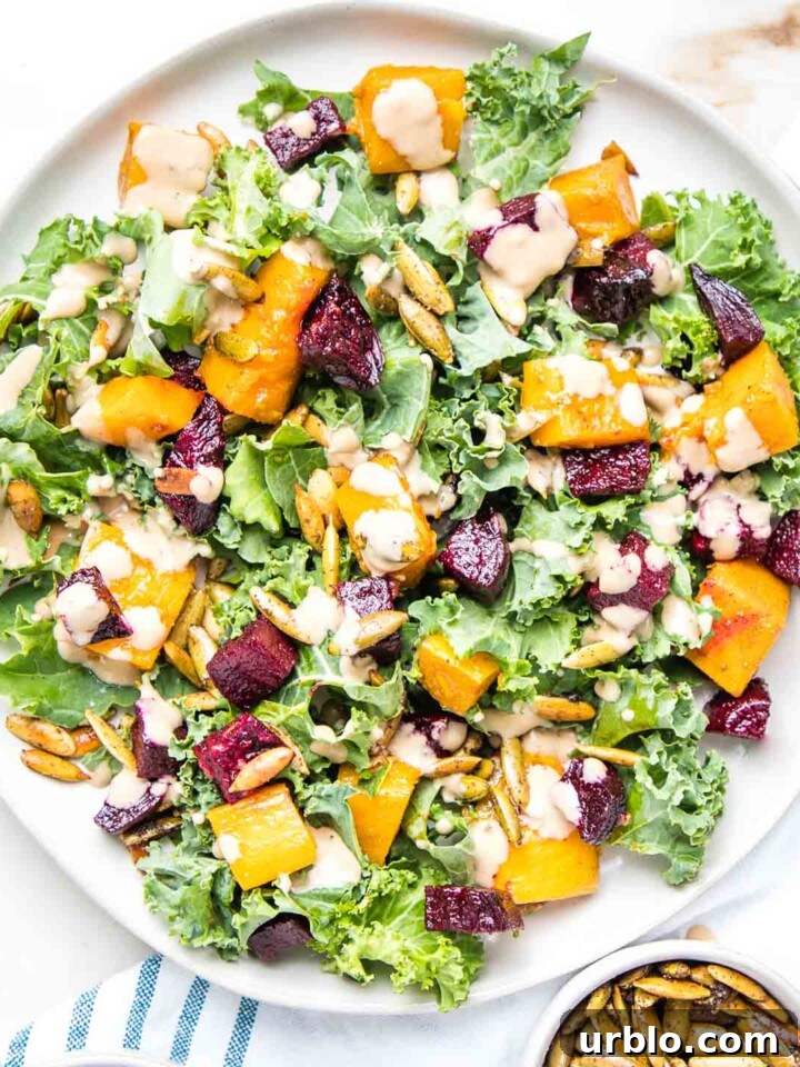 Large rimmed white plate filled with kale, roasted butternut squash and beets, toasted pumpkin seeds and a creamy balsamic dressing on top of a white tea towel with blue stripes and a small dish of extra toasted pumpkin seeds.