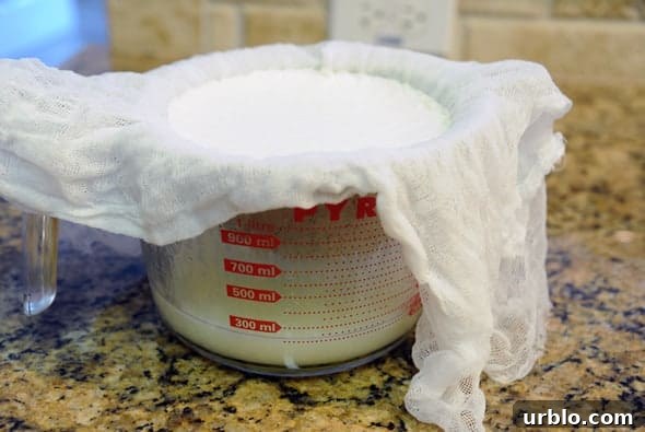 boiled milk resting with in a large glass measuring cup topped with a cheese cloth