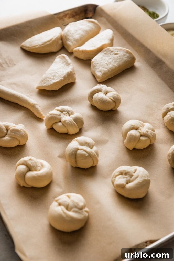 Large sheet pan lined with parchment paper and filled with garlic knot dough in various stages of tying and twisting.