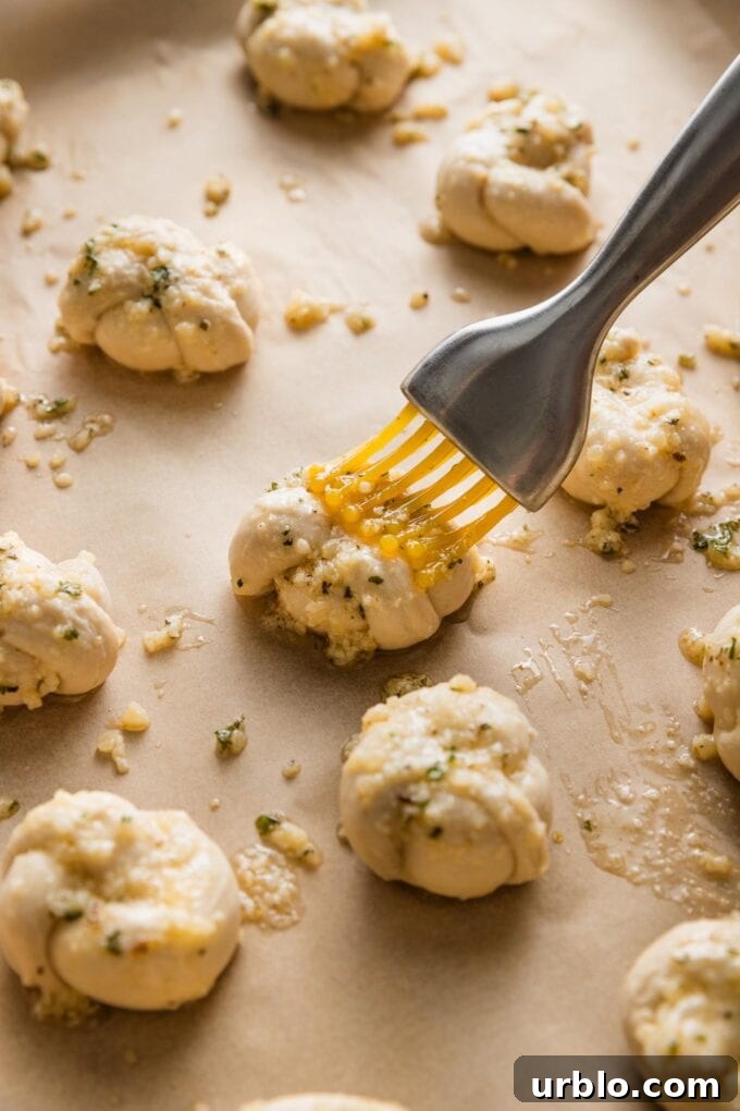 Close up view of a pastry brush coating small, unbaked garlic knots with a butter Parmesan herb mixture.