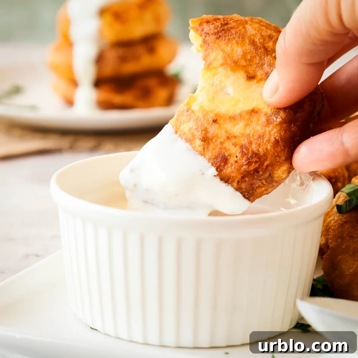 Crispy Potato Fritters Recipe for Golden, Crunchy Hash Browns 9 Side shot of a hand dipping a potato fritter in cheese sauce in a ramekin with more fritters in the background.