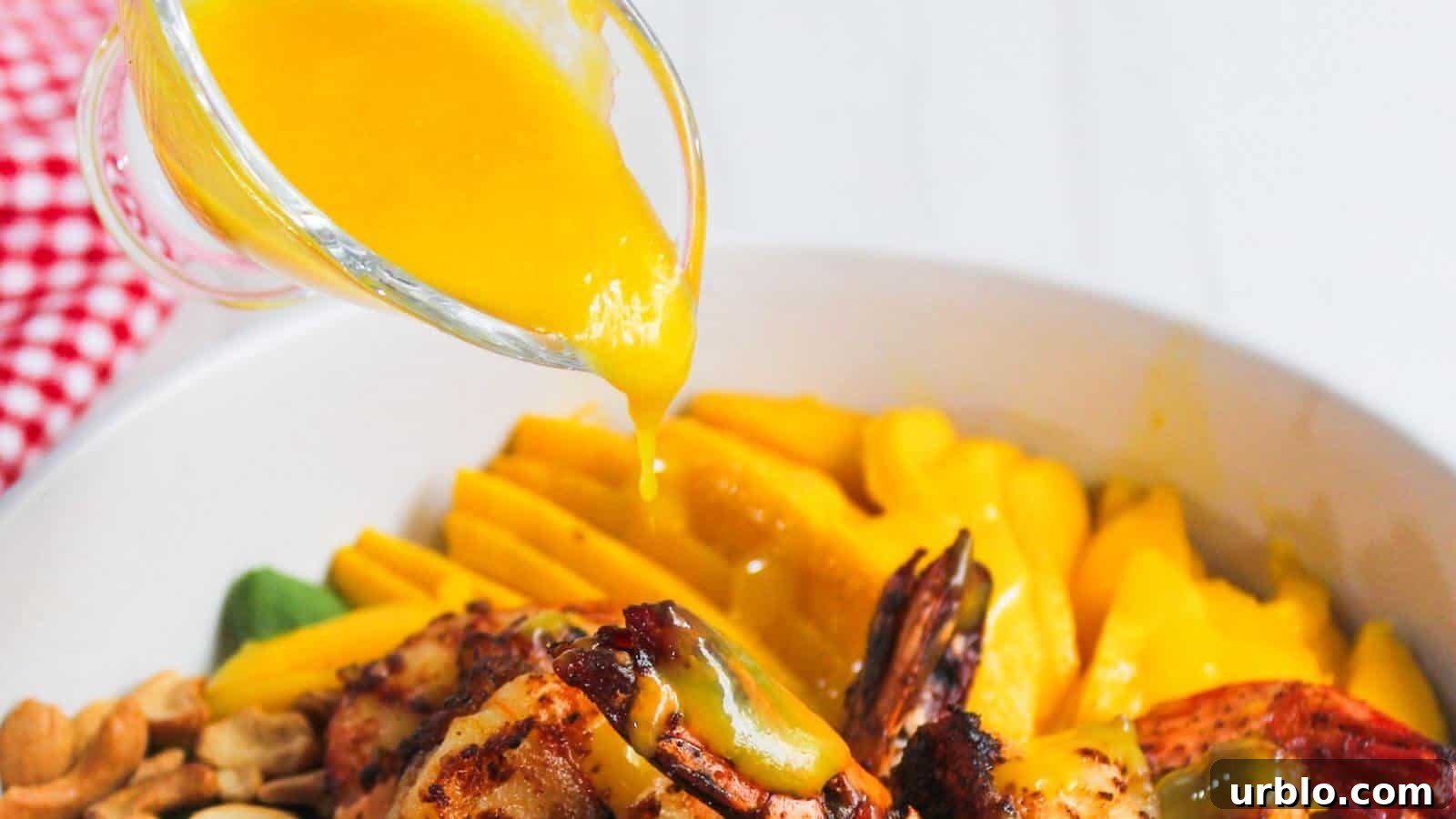 A glass container pours yellow sauce over grilled shrimp in a bowl with mango slices, nuts, and leafy greens.