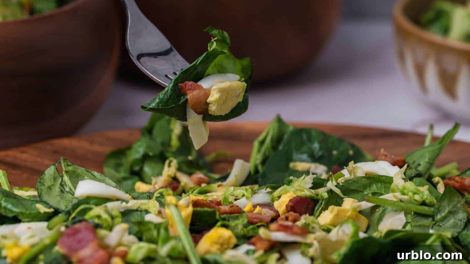 A fork holds a bite of spinach salad with chopped eggs, bacon, and cheese over a wooden bowl filled with the same salad. Two blurred bowls are in the background.