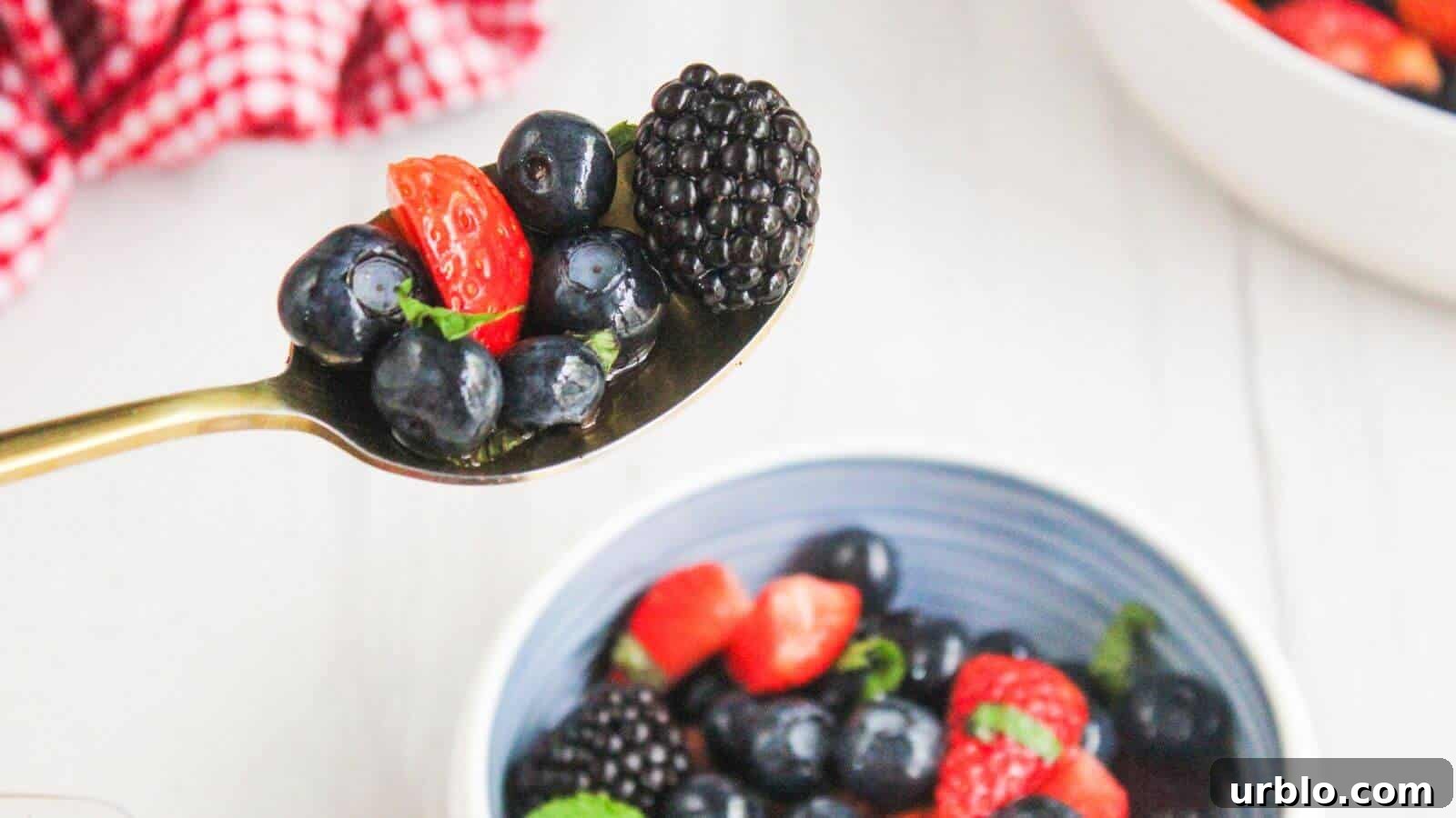 A spoon holding blueberries, a sliced strawberry, and a blackberry above a bowl of mixed berries on a white surface.