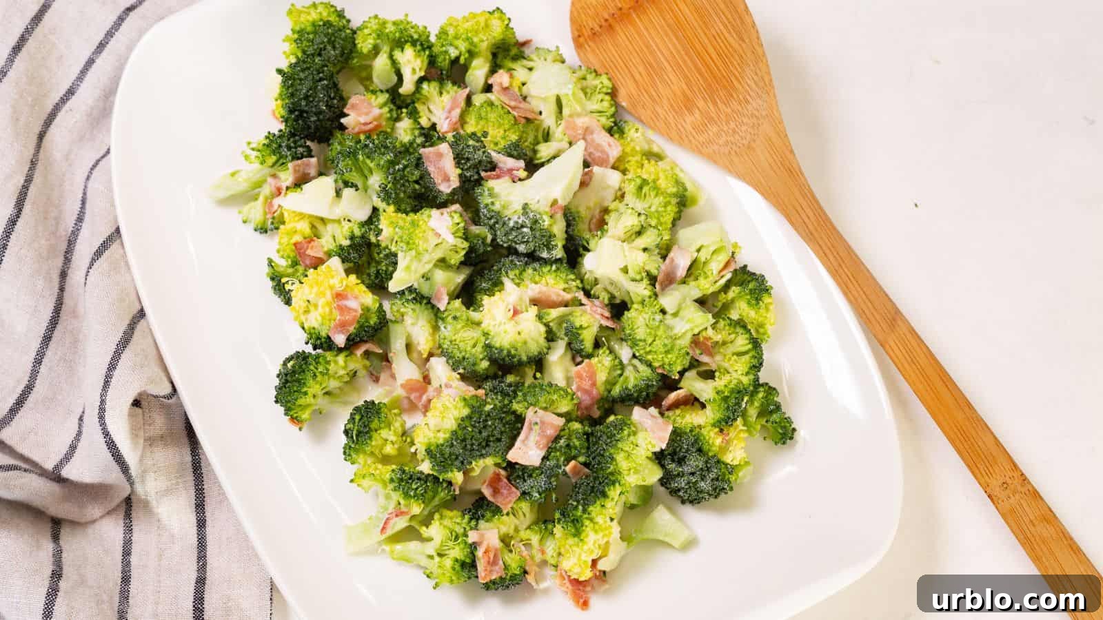 Broccoli salad with bacon bits on a white oval plate, accompanied by a wooden serving spoon on a linen napkin.