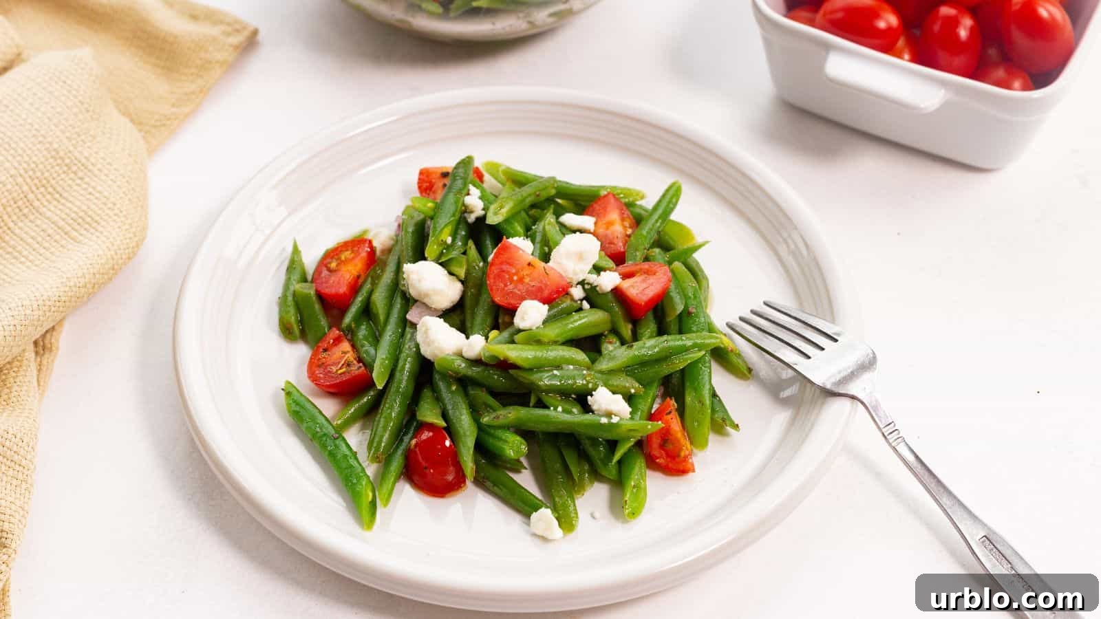 A plate of green beans tossed with cherry tomatoes and crumbled feta cheese, accompanied by a fork, on a white surface.