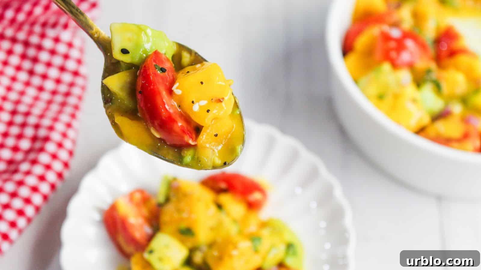 A spoonful of colorful mango avocado salad is held above a white plate, with a bowl of the same vibrant salad in the background.