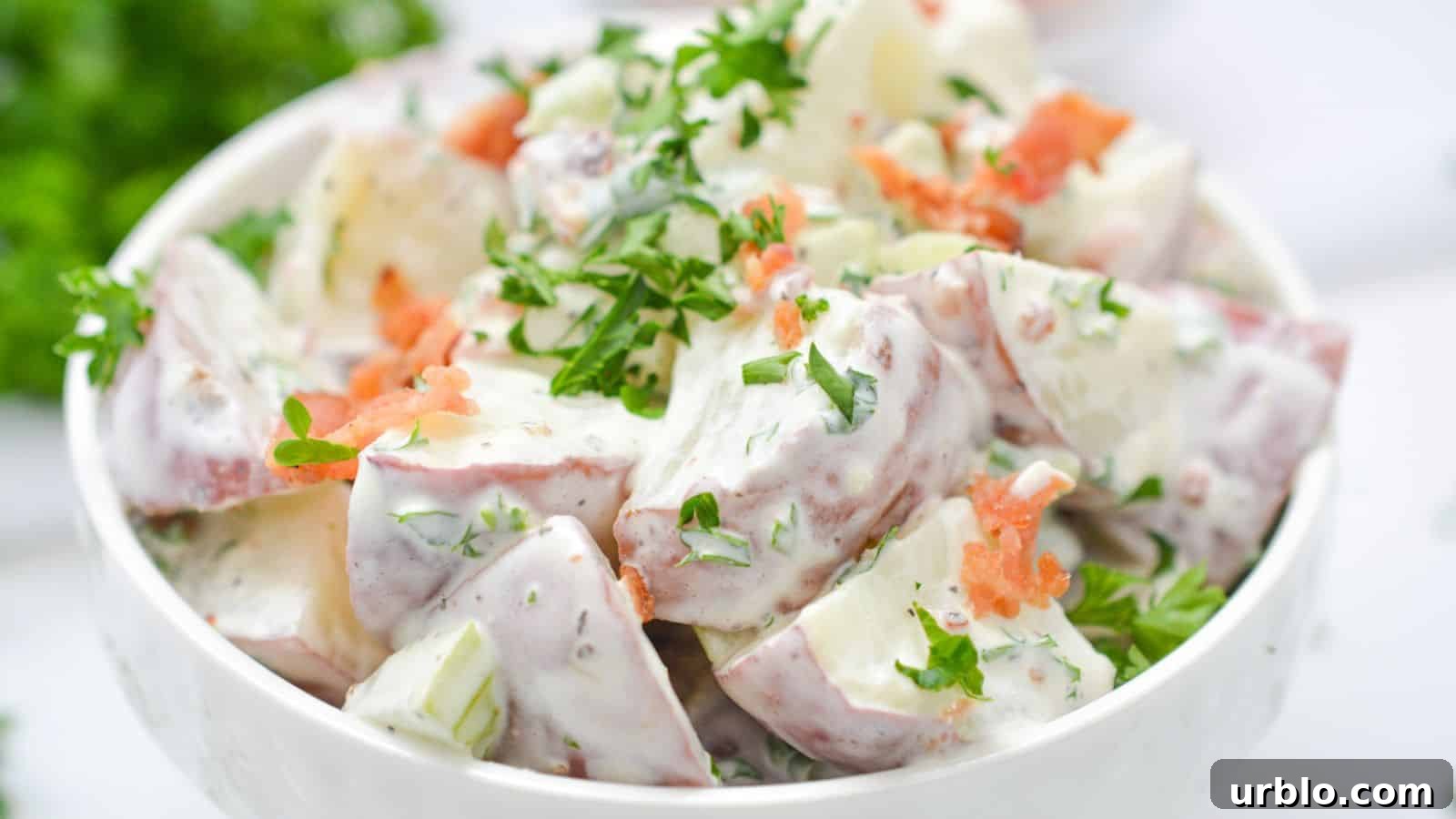 Bowl of potato salad garnished with fresh herbs and bacon on a white background.