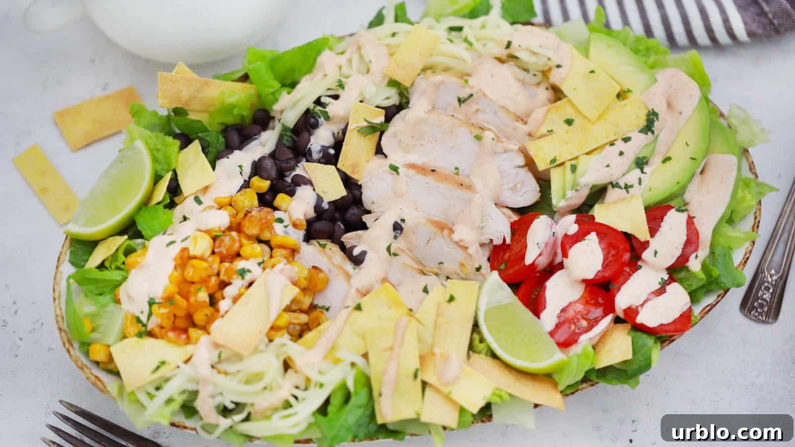A colorful chicken salad with avocado, black beans, corn, cherry tomatoes, and lime slices, presented in a white bowl.