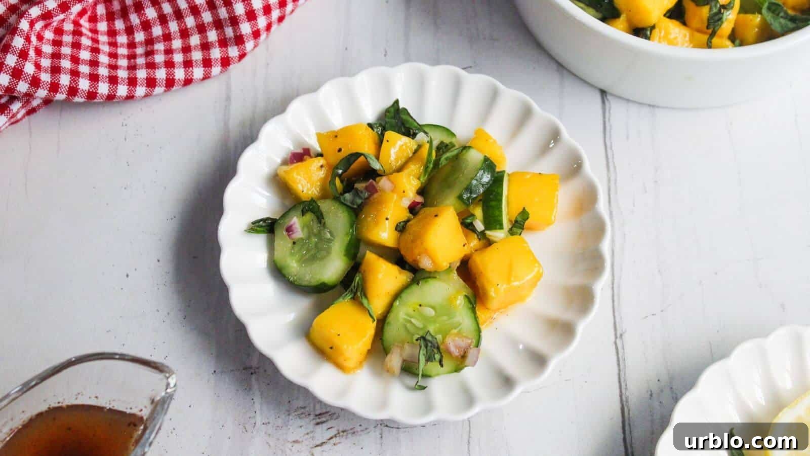 A white plate containing a salad with cucumber slices, mango chunks, red onion, and fresh herbs sits on a white surface.