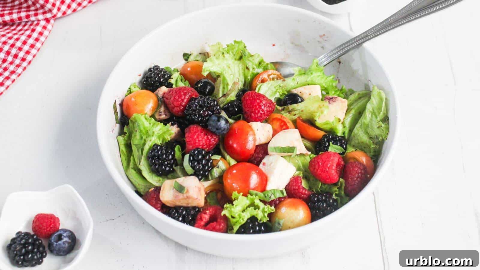 A white bowl filled with leafy salad, cherry tomatoes, berries, avocado, and a metal spoon, with a small dish of dark dressing and a red checkered cloth nearby.