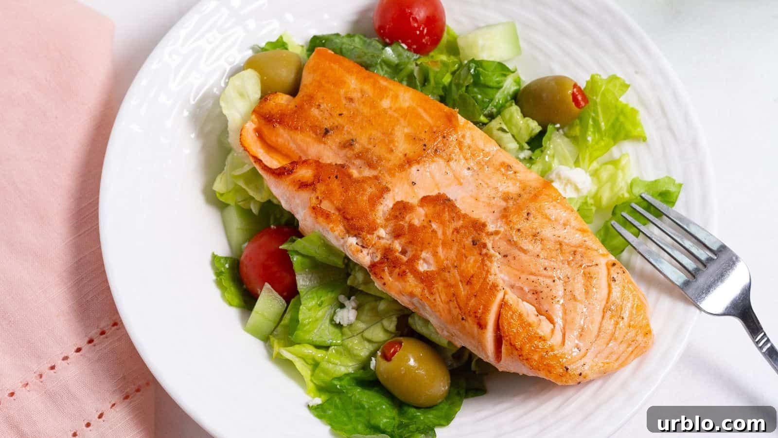 A grilled salmon fillet served on a bed of fresh salad with lettuce, tomatoes, and olives on a white plate.
