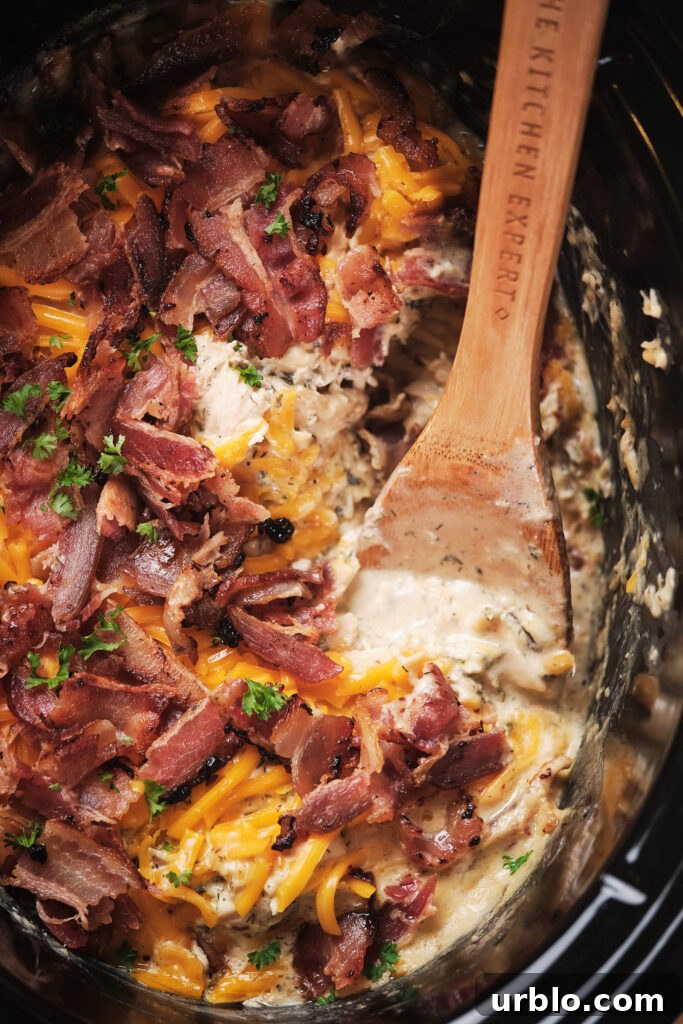 Close-up of a creamy chicken and cheese dish topped with crispy bacon and parsley in a slow cooker, with a wooden spoon.