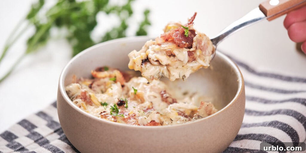 A spoon scooping creamy chicken casserole with bacon and parsley from a bowl placed on a striped cloth.