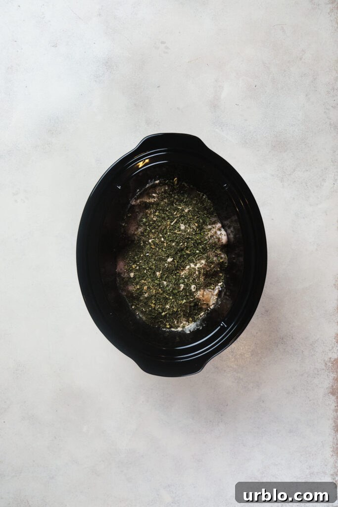 A black crock pot with chicken and herbs on a light grey surface.