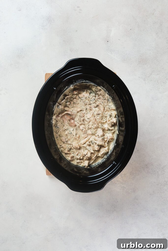 A creamy chicken mixture in a black crock pot on a light grey surface.