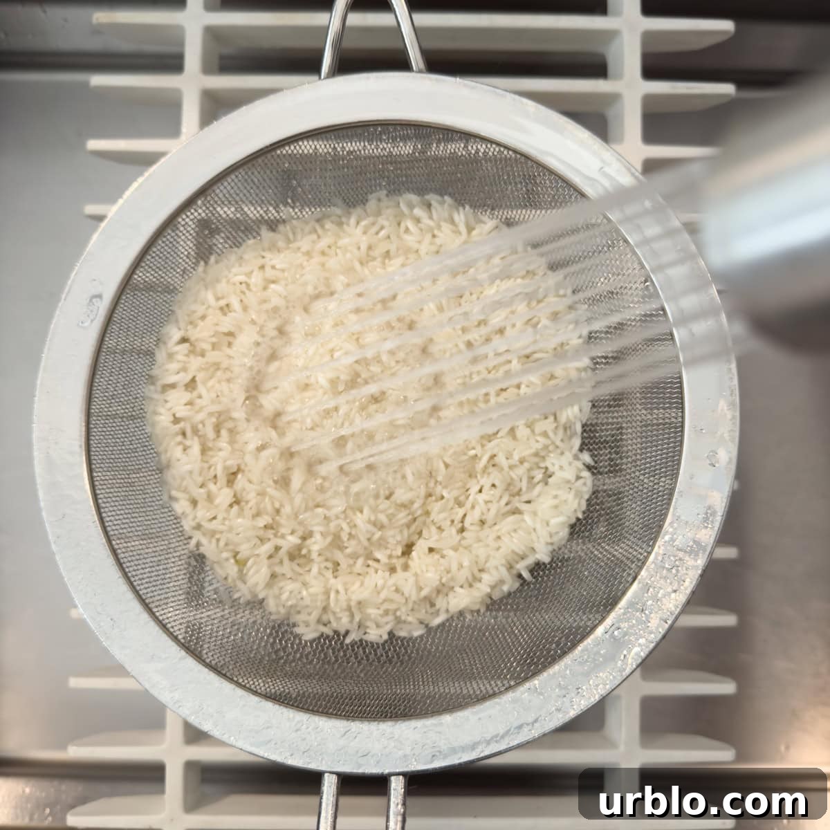 White rice in a sieve being washed in the sink with a sprayer.