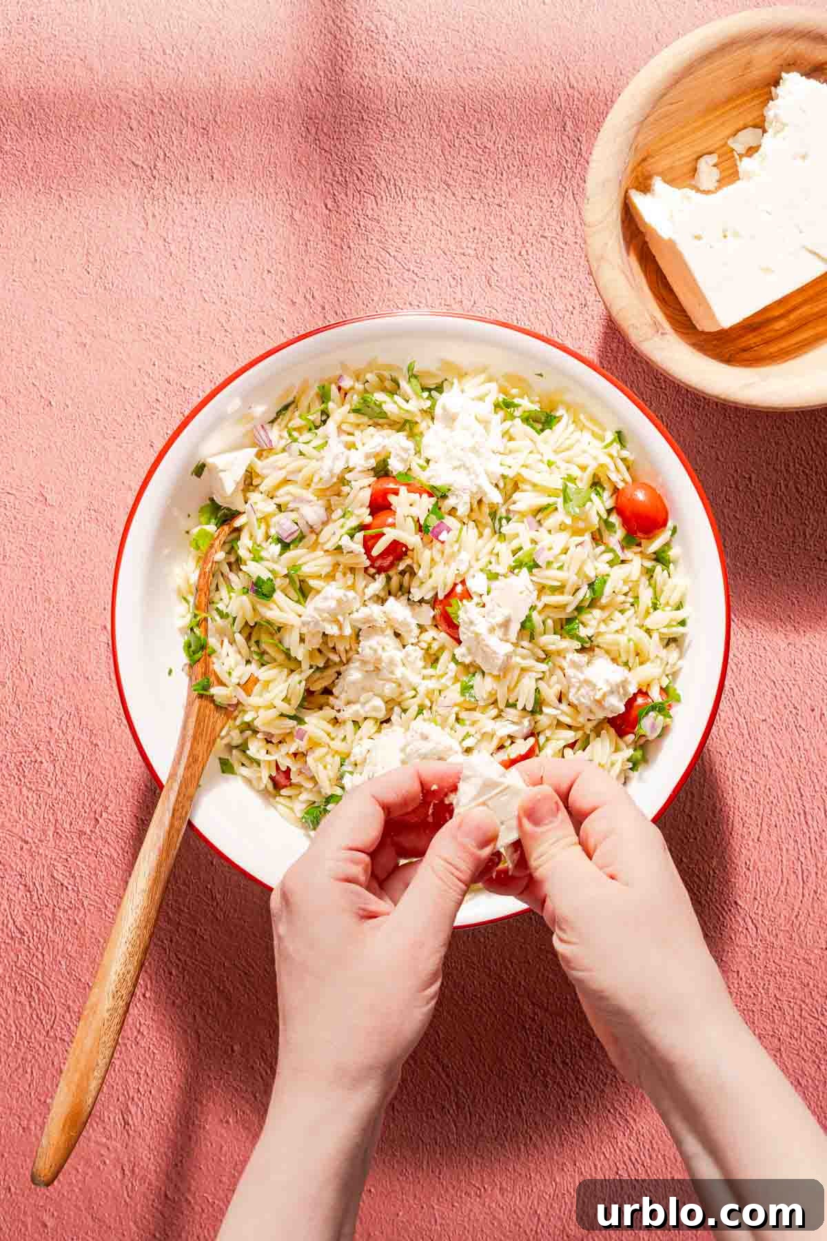 hands crumbling feta cheese over orzo salad with bowl of feta on the side.