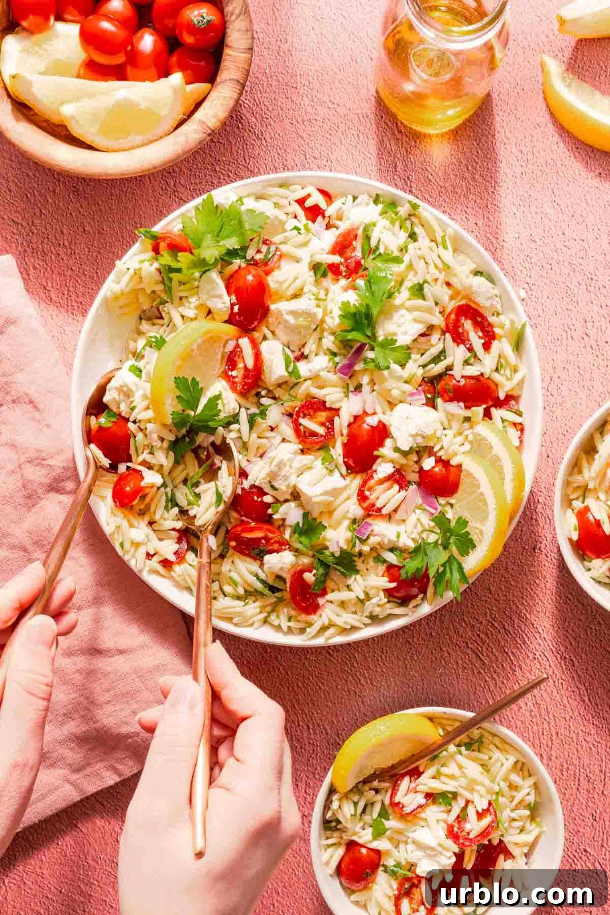 lemon orzo salad in white bowl with hands lifting serving out with two spoons.