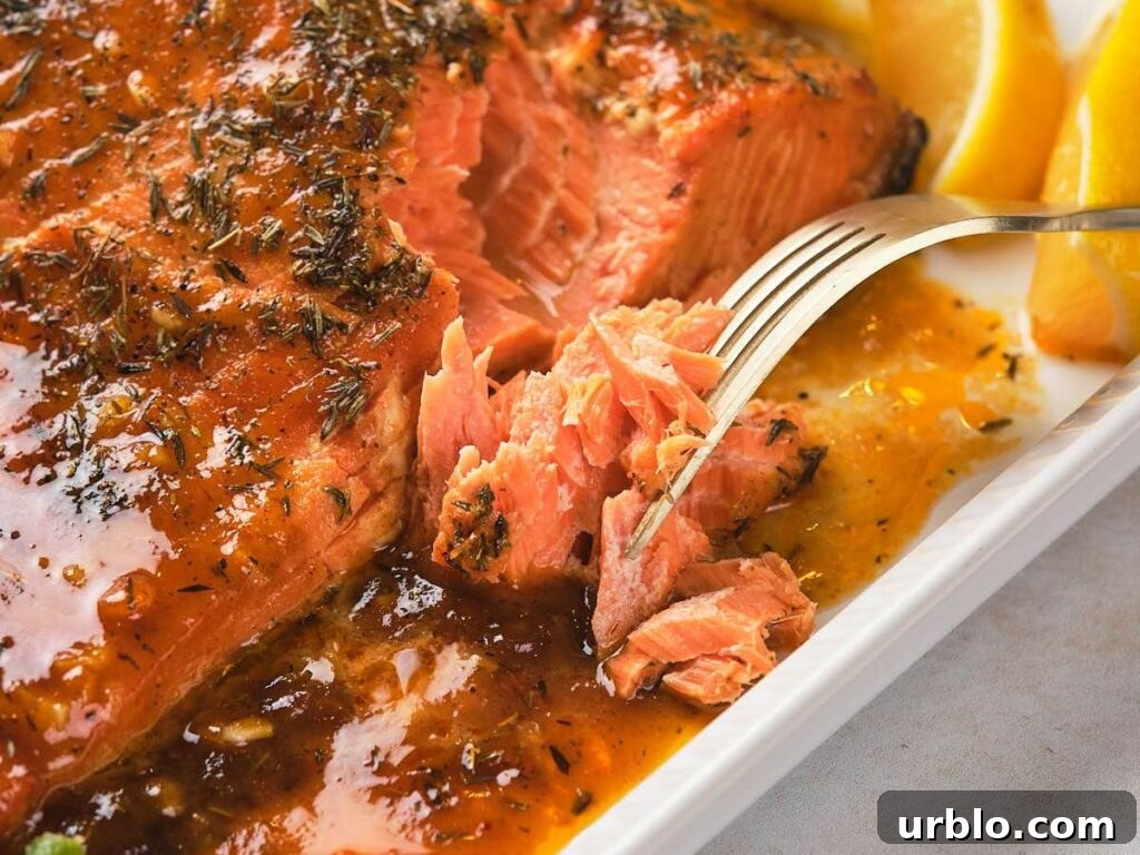 Fork breaking into a glazed, baked salmon fillet garnished with herbs and accompanied by lemon wedges on a white plate.