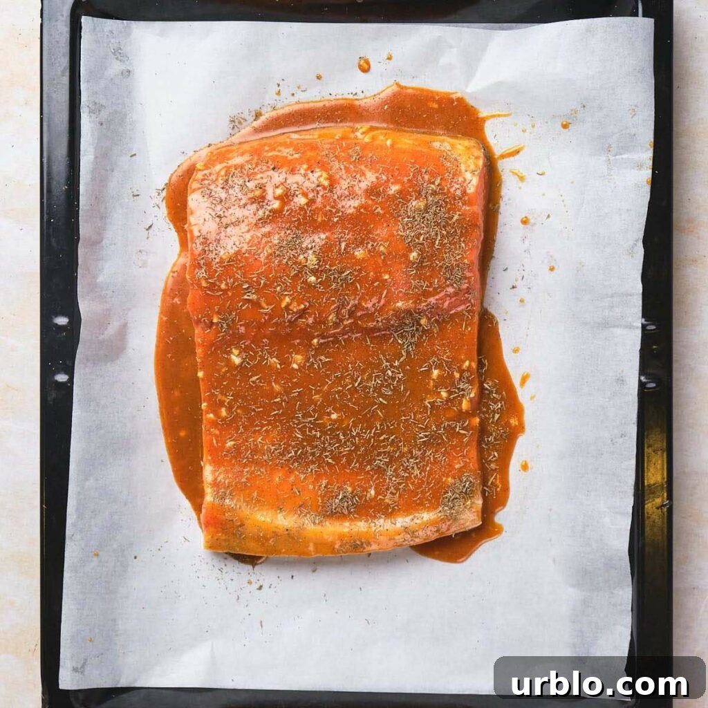 Salmon fillet coated in a red sauce and sprinkled with herbs on a baking sheet lined with parchment paper.