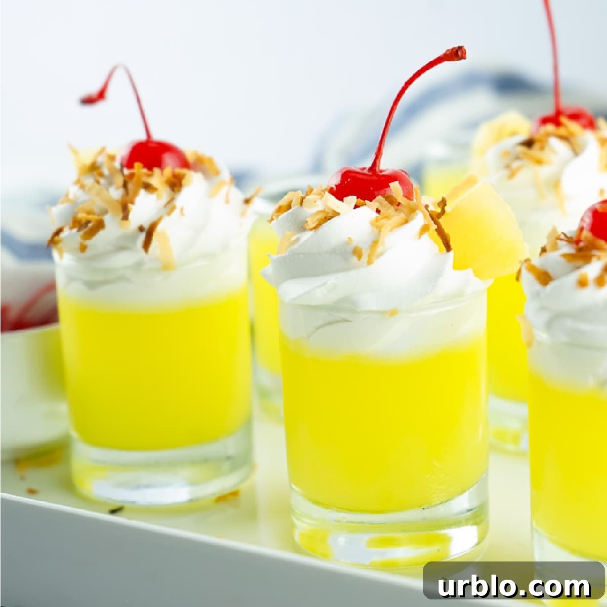 Pina Colada Jello Shots topped with whipped cream, toasted coconut, and cherries on a white tray.