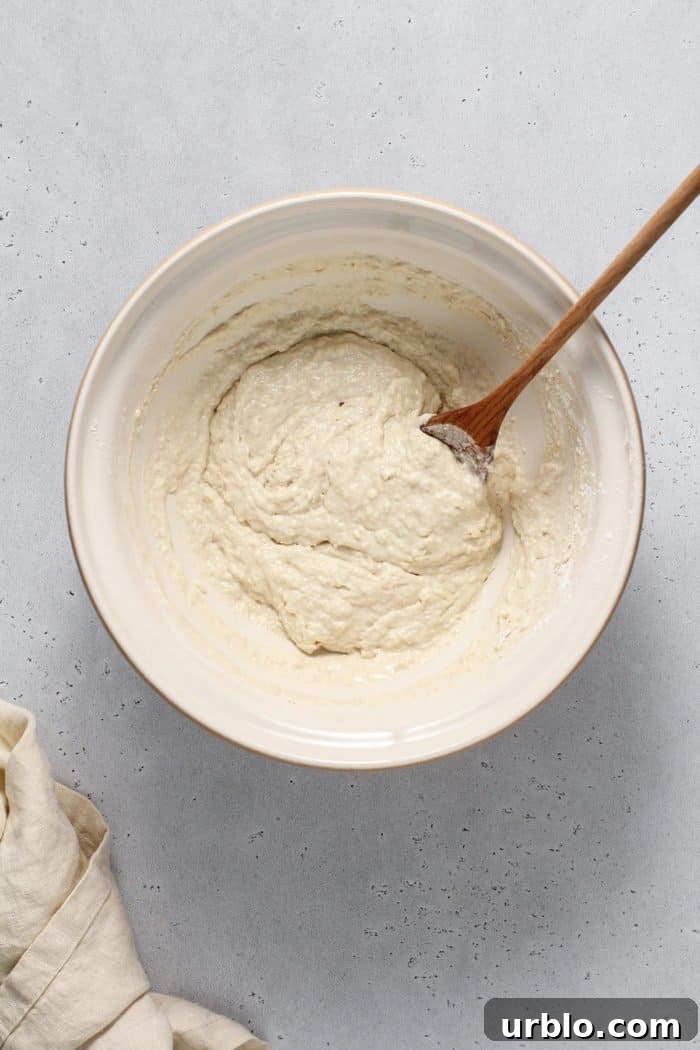 English muffin bread dough in a white mixing bowl, being stirred with a wooden spoon