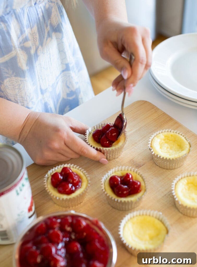 Mini Cherry Cheesecakes Made in a Muffin Tin 8 Person spooning cherry topping onto mini cheesecakes on a wooden board.