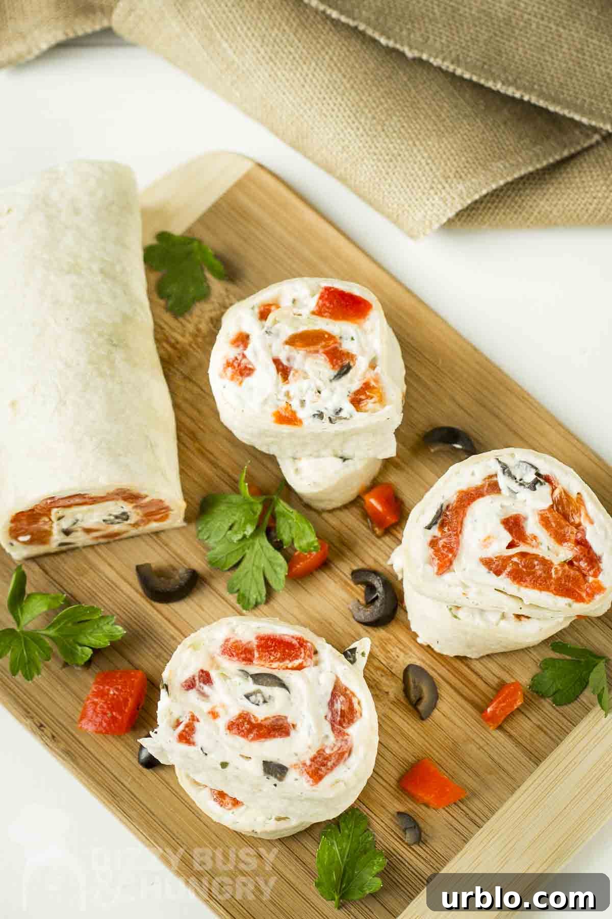 Creamy Ranch Pinwheels with Roasted Red Peppers Appetizer 2 Overhead shot of three pinwheels cut from the pinwheel log on a wooden cutting board with sliced olives, herbs, and red peppers scattered around.