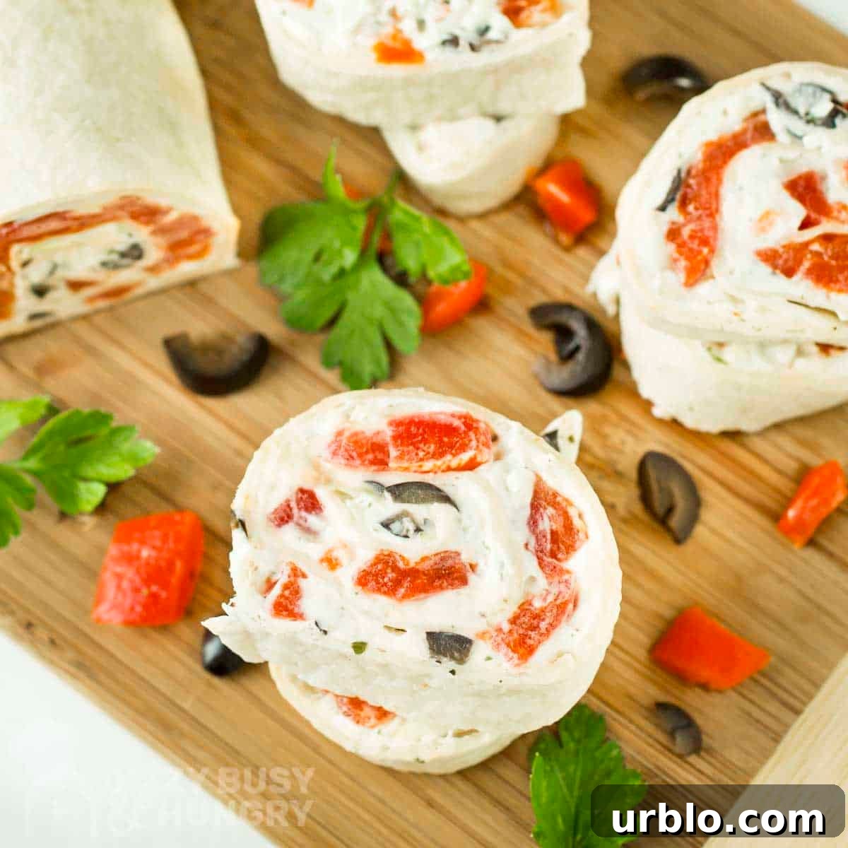Creamy Ranch Pinwheels with Roasted Red Peppers Appetizer 11 Overhead shot of three pinwheels cut from the pinwheel log on a wooden cutting board with sliced olives, herbs, and red peppers scattered around.