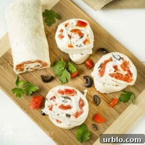 Creamy Ranch Pinwheels with Roasted Red Peppers Appetizer 12 Overhead shot of three pinwheels cut from the pinwheel log on a wooden cutting board with sliced olives, herbs, and red peppers scattered around.