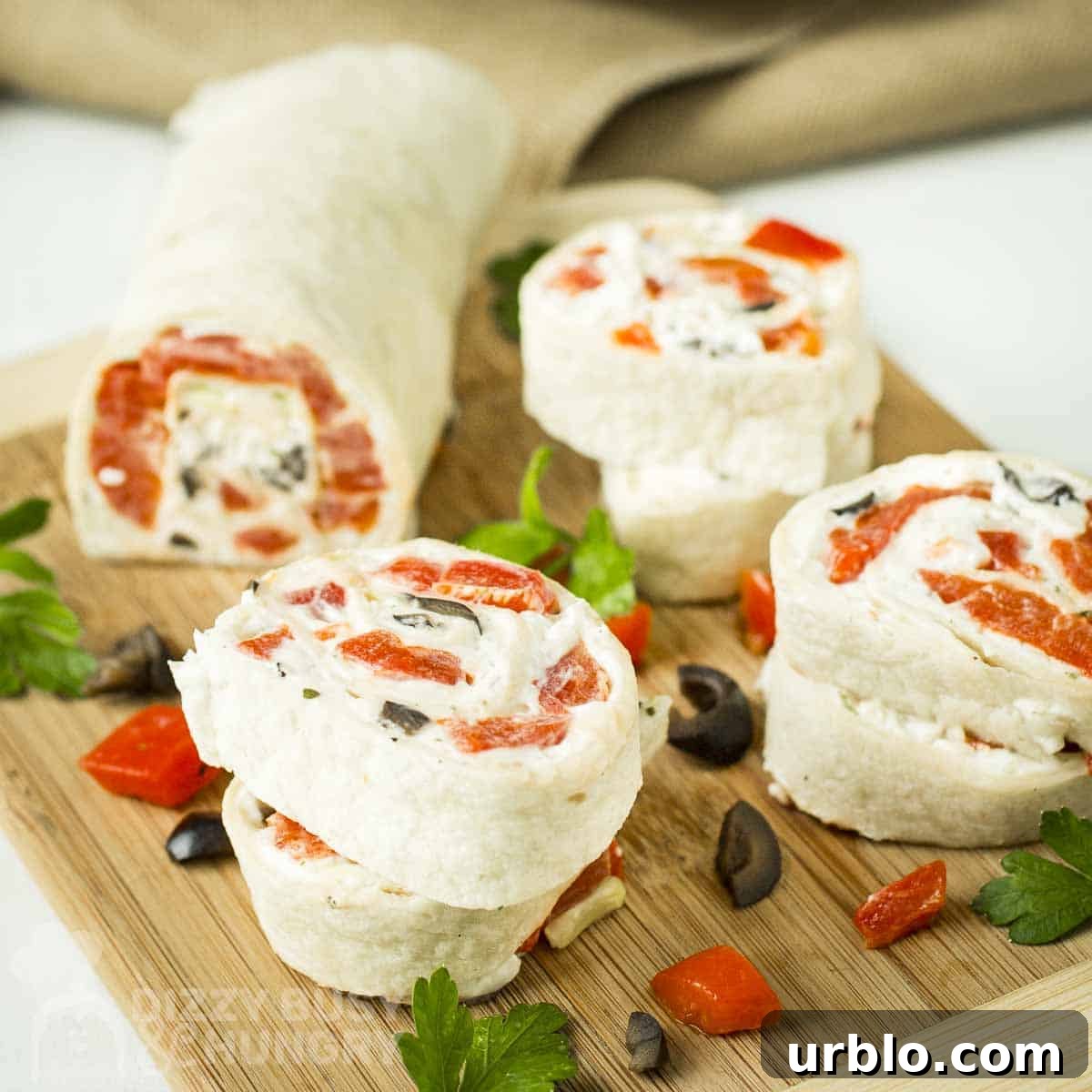 Creamy Ranch Pinwheels with Roasted Red Peppers Appetizer 10 Side shot of multiple pinwheels cut from the pinwheel log on a wooden cutting board with sliced olives, herbs, and red peppers scattered around.